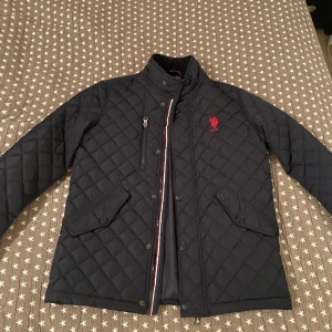 Mörkblå quiltad jacka från U.S. Polo Assn. - Snygg mörkblå quiltad jacka från U.S. Polo Assn. med röd broderad logga på bröstet. Jackan har en dragkedja och knappar framtill samt praktiska fickor. Insidan har ett stilrent rutigt mönster i rött, svart och vitt.