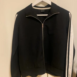 Svart bomberjacka med vita ränder - Snygg svart bomberjacka med vita ränder längs ärmarna. Jackan har dragkedja framtill och en klassisk krage. Perfekt för en sportig look.