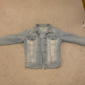Ljusblå jeansjacka - Säljer en stilren ljusblå jeansjacka med klassisk krage och knappar framtill. Jackan har två bröstfickor med knappdetaljer och är perfekt för en avslappnad look.