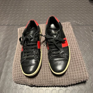 Saint Laurent - Snygga svarta sneakers med röda detaljer från Saint Laurent. Skorna har snörning och en klassisk design med en kontrasterande vit sula. Perfekta för en stilren look.