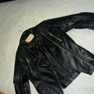 Svart skinnjacka från Pepellas - Snygg svart skinnjacka från voyelles  med dragkedjor och quiltade detaljer på axlar och ärmar. Perfekt för en cool look. Jackan har en klassisk bikerstil med flera fickor och en bekväm passform.