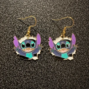 Färgglada örhängen med Stitch - Säljer ett par unika örhängen med en glad tecknad figur i blått, lila och svart. De har guldfärgade krokar och är perfekta för att ge en lekfull touch till din stil.