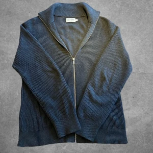 Mörkgrå ribbad kofta från East West - Säljer en stilren mörkgrå kofta från East West i 100% bomull. Koftan har en dragkedja framtill och är ribbad för en snygg textur. Perfekt för kyligare dagar.