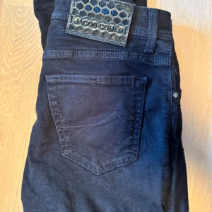 Svarta jeans från Jacob Cohen - Snygga svarta jeans från Jacob Cohen med knappgylf och klassisk femficksdesign. Tillverkade i Italien med högkvalitativt material för en stilren look. Perfekta för dig som vill ha en tidlös stil. Nypris 5000kr säljer för 1245kr