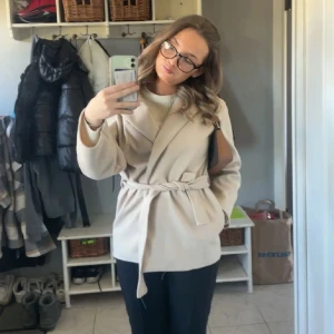 Beige kappa med knytband - Snygg beige kappa med knytband i midjan. Perfekt för höst och vår. Den har en stilren design med långa ärmar och en mjuk, bekväm passform.