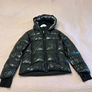 Moncler jacka  - Snygg svart pufferjacka från Moncler med glansig finish och logotyp på huvan. Jackan har en dragkedja framtill och långa ärmar med ribbade muddar. Perfekt för kyliga dagar.