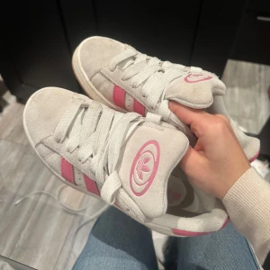 Adidas Campus sneakers i vitt/grå och rosa - Snygga Adidas Campus sneakers i vitt med rosa detaljer. Skorna har klassiska tre ränder på sidan och en bekväm sula. Perfekta för en avslappnad stil. Skorna är lite använda dock är dem i väldigt bra skick. 