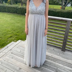 Silverfärgad långklänning med paljetter - Elegant långklänning i silver med glittrande paljetter upptill och en v-ringad design. Klänningen har en luftig kjol i ett lätt material som ger en vacker fall. 