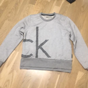 Grå sweatshirt från Calvin Klein Jeans - Säljer en stilren grå sweatshirt från Calvin Klein Jeans med det ikoniska CK-logotypen i svart på framsidan. Tröjan har långa ärmar och ribbade muddar vid ärmslut och nederkant. Perfekt för en avslappnad look.