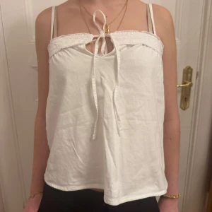 Vit topp med spetsdetaljer - Säljer en söt vit topp med smala axelband och spetsdetaljer upptill. Toppen har en knytning framtill och är helt ny.