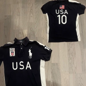 USA Polo Ralph Lauren - Säljer en svart pikétröja från Polo Ralph Lauren med USA-tryck och nummer 10 på ryggen. Tröjan har en klassisk krage och korta ärmar med vita detaljer på sidorna. Perfekt för en sportig look!