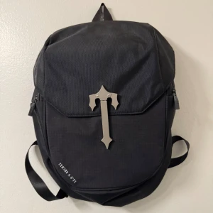 Trapstar ryggsäck - Snygg svart ryggsäck från Trapstar med ett unikt metallkors på framsidan. Ryggsäcken har justerbara axelremmar och en praktisk dragkedja. Perfekt för att bära dina saker med stil.