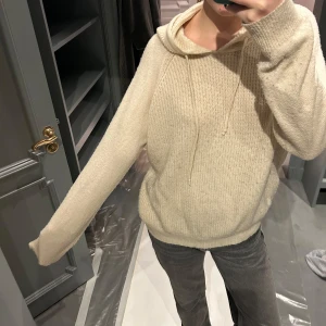 Beige stickad hoodie - Mysig beige stickad hoodie med luva och långa ärmar. Perfekt för kyliga dagar med en avslappnad passform. Den har en klassisk design med ribbade muddar vid ärmslut och nederkant.