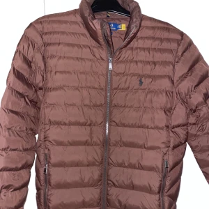 Polo Ralph Lauren Brun pufferjacka - Säljer en snygg brun pufferjacka från Polo Ralph Lauren. Jackan har en dragkedja framtill och två praktiska fickor med dragkedjor. Perfekt till våren, endast använd ett fåtal gånger så den är som ny!