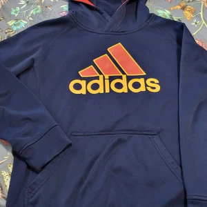 Mörkblå hoodie från Adidas - Säljer en snygg mörkblå hoodie från Adidas med röd och gul logga på framsidan. Hoodien har en bekväm passform och en praktisk huva. Hund finns hemma.