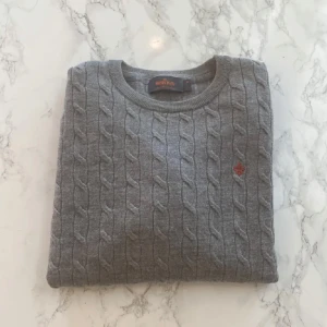 Grå kabelstickad tröja från Morris - Säljer en stilren grå kabelstickad tröja från Morris i 100% merinoull. Tröjan har en klassisk rund hals och ett diskret broderat emblem på bröstet. Perfekt för kyligare dagar.