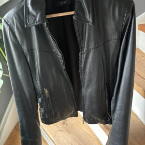 Svart skinnjacka från Pour - Snygg svart skinnjacka från Pour med dragkedja framtill och på ärmsluten. Jackan har en klassisk krage och två fickor med dragkedja. Perfekt för en cool och stilren look.