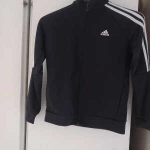 Svart träningsjacka från Adidas - Snygg svart träningsjacka från Adidas med klassiska vita ränder på ärmarna. Jackan har en dragkedja framtill och en hög krage. Perfekt för sport och träning.