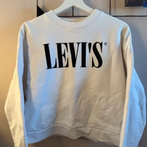 Vit sweatshirt från Levi's - Säljer en vit sweatshirt från Levi's med deras ikoniska logga tryckt i svart på framsidan. Tröjan har långa ärmar och en rund halsringning. 