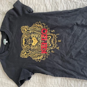 Svart t-shirt med tigertryck från Kenzo - Snygg svart t-shirt från Kenzo med ett coolt tigertryck i guld och rött på framsidan. Perfekt för att ge din outfit en edgy touch. Kortärmad och tillverkad i mjukt material för bekväm passform. Storlek XS!