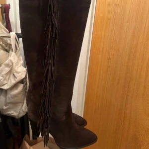 Bruna mockastövlar med fransar - Snygga bruna stövlar i mocka från Zara med fransdetaljer längs sidan. De har en spetsig tå och en stabil klack som ger en elegant look. Perfekta för att ge din outfit en bohemisk touch.