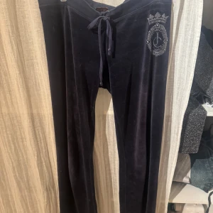 Mörkblå velourbyxor från Juicy Couture - Säljer ett par mörkblå velourbyxor från Juicy Couture med en snygg broderad logga på höften. Byxorna har en bekväm passform med snörning i midjan för justerbarhet. Perfekta för en avslappnad och stilren look.