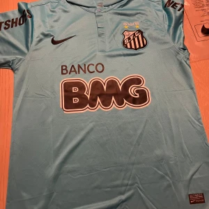 Ljusblå fotbollströja från Nike - Säljer en ljusblå fotbollströja från Nike med Santos FC-logga och sponsortryck. Tröjan har korta ärmar och en klassisk krage. Perfekt för fotbollsfans som vill ha en unik tröja i sin samling.