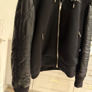 Svart bomberjacka från Givenchy med läder på armarna - Snygg svart bomberjacka från Givenchy med quiltade läderärmar och dragkedja framtill. Jackan har en stilren design med ribbade muddar och Givenchy-logga på bröst.  Nypris 30.000kr. Kvitto medföljer, inköpt i butik i London!