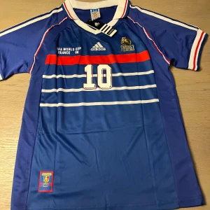 Blå fotbollströja från Adidas - Säljer en blå fotbollströja från Adidas med korta ärmar och vita samt röda detaljer. Tröjan har nummer 10 och texten 'Zidane' på ryggen. Perfekt för fotbollsfans!