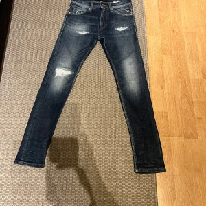 Mörkblå jeans från Replay - Snygga mörkblå jeans från Replay med slitna detaljer och en klassisk femficksdesign. De har en läderpatch med Replay-logga på baksidan och en dragkedja i gylfen. Perfekta för en avslappnad stil.