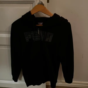 Svart hoodie från Puma - Säljer en svart hoodie från Puma med dragkedja och stort tryck på framsidan. Perfekt för en avslappnad stil. Den har en justerbar huva och långa ärmar.