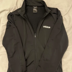 Svart kofta från Adidas - Säljer en svart kofta från Adidas med dragkedja och hög krage. Jackan har en stilren design med Adidas-loggan på bröstet. Perfekt för träning eller som en lätt överdragströja. 🖤