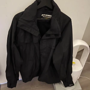 Svart vindjacka från Everest - Säljer en svart vindjacka från Everest. Jackan har en stilren design med dragkedja framtill och långa ärmar. Perfekt för blåsiga dagar och passar bra till vardagsäventyr. En klassisk och funktionell jacka för alla tillfällen.