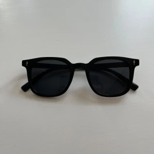 Svarta solglasögon med fyrkantig form - Snygga svarta solglasögon med en klassisk fyrkantig form. De har en stilren design med mörka linser och små detaljer på bågarna. 