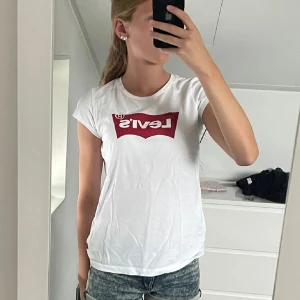 Vit t-shirt från Levi's - Säljer en klassisk vit t-shirt från Levi's med det ikoniska röda logotyptrycket på bröstet. T-shirten har en rund halsringning och korta ärmar. Perfekt för en avslappnad stil.