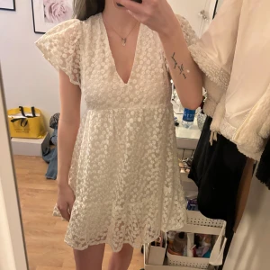 Vit blommig klänning - Charmig vit klänning med blommigt mönster och v-ringad design. Klänningen har korta ärmar med volangdetaljer och en öppen rygg. Perfekt för studenten eller i sommar !!! Säljer då jag hittat en annan klänning jag ska ha på studenten. Undershorts inbyggda i klänningen 💕