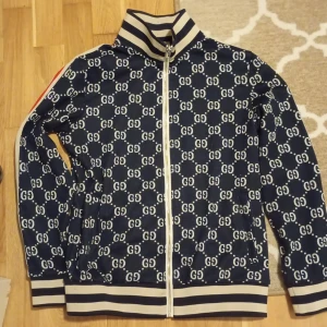 Blå mönstrad tröja från Gucci - Säljer en snygg blå jacka från Gucci med ett ikoniskt GG-mönster. Jackan har en dragkedja framtill och ribbade muddar. Den har också röda och vita ränder längs ärmarna för en sportig look.