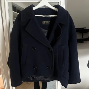 Ull kappa Zara - Snygg mörkblå kappa i ull från Zara med dubbelknäppning och bred krage. Jackan har en klassisk design med två framfickor och är perfekt för kyligare dagar. Tillverkad i samarbete med Manteco för en lyxig känsla. Nypris 1395kr