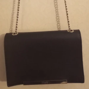 Svart axelväska från KappAhl - Snygg svart axelväska från KappAhl med kedjerem och stilren design. Väskan har en praktisk dragkedja inuti och en elegant stängning med dekorativa detaljer. Perfekt för att bära dina nödvändigheter med stil.