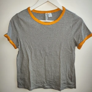 Randig t-shirt med gula detaljer - Säljer en randig t-shirt från H&M med gula kanter vid hals och ärmslut. T-shirten har korta ärmar och en klassisk rund halsringning. Perfekt för en avslappnad stil.