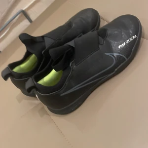 Svarta Nike Air Zoom fotbollsskor - Snygga svarta Nike Air Zoom fotbollsskor med en stilren design. Skorna har en elastisk rem över vristen för extra stöd och en slitstark sula med bra grepp. Perfekta för fotbollsplanen med en modern look.