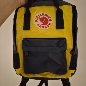 Gul och blå ryggsäck från Fjällräven - Säljer en klassisk Fjällräven Kånken Mini ryggsäck i en snygg gul och blå färgkombination. Ryggsäcken har ett huvudfack och en praktisk framficka med dragkedja. Onamnad och sittunderlag finns med.Perfekt för både skola och utflykter! Finns någon liten fläck, se bild.