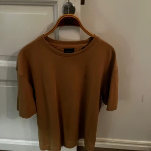 Brun kortärmad tröja från GANT - Säljer en stilren brun kortärmad tröja från GANT. Tröjan har en ribbad krage och en diskret logga på bröstet. Perfekt för en avslappnad stil.