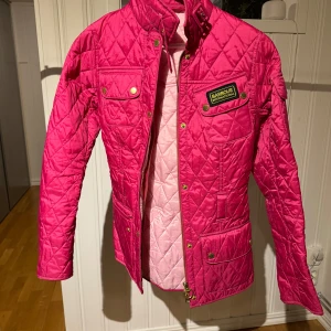 Rosa quiltad jacka från Barbour - Säljer en snygg rosa quiltad jacka från Barbour. Jackan har en klassisk design med knappar och dragkedja framtill samt en stilren krage. Perfekt för att ge lite färg till din garderob!