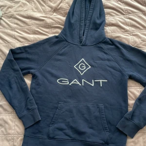 Mörkblå hoodie från GANT - Säljer en snygg mörkblå hoodie från GANT med stor logga på bröstet. Tröjan har en klassisk känguruficka och en bekväm huva. Perfekt för en avslappnad stil.