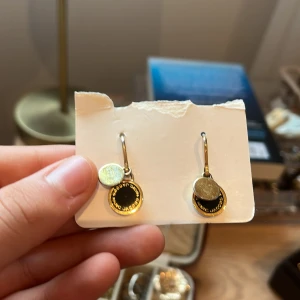 Guldörhängen med svart och silverdetaljer - Snygga guldörhängen med rund svart detalj och en mindre silverfärgad platta. Perfekta för att ge en elegant touch till din outfit. De har en enkel krok för enkel användning.