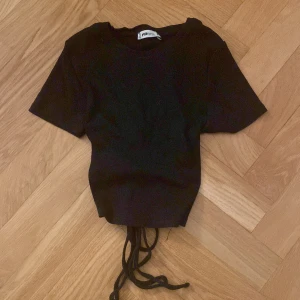 Svart topp med snörning från FB Sister - Snygg svart topp från FB Sister med korta ärmar och cool snörning i ryggen. Perfekt för en trendig look. Tillverkad i ett mjukt material som ger en skön passform.