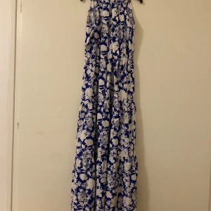 Blå och vit blommig långklänning - Säljer en vacker långklänning med ett blommigt mönster i blått och vitt. Klänningen har en elegant design som passar perfekt för sommarens alla tillfällen. Den är ärmlös och har en luftig passform som ger en skön känsla. Perfekt för dig som vill ha en stilren och bekväm look.