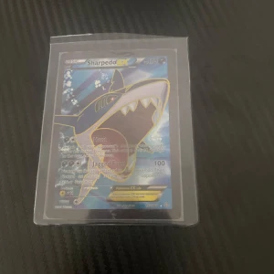 Sharpedo EX Pokémonkort - Säljer ett Sharpedo EX Pokémonkort med en cool design. Kortet har en bild av Sharpedo med en blå bakgrund och är inneslutet i ett plastfodral för extra skydd. Perfekt för samlare eller Pokémon-fans!