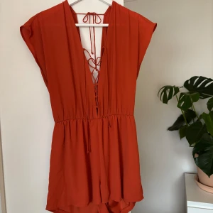 Orange byxdress med snörning - Snygg orange byxdress med korta ärmar och v-ringad design. Den har en unik snörning i ryggen som ger en extra detalj. Perfekt för sommarens varma dagar.